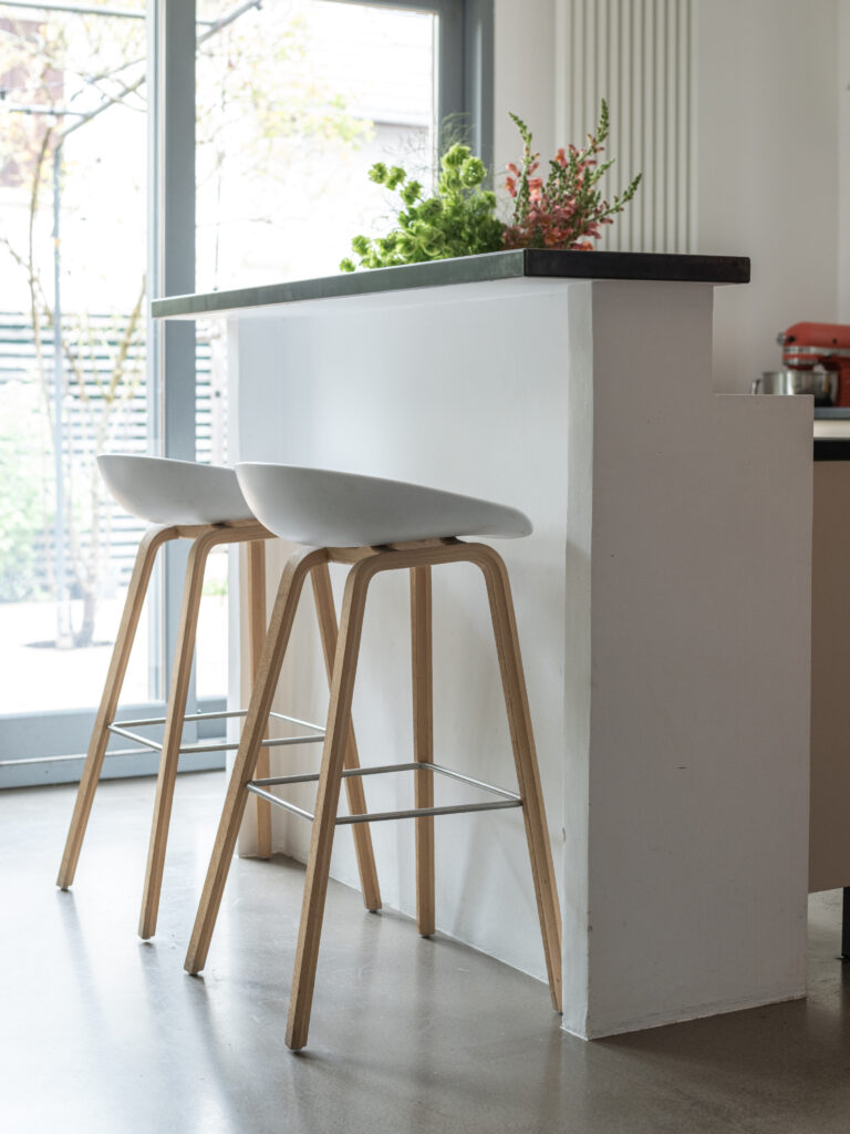 Deux tabourets en bois clair avec des sièges blancs dans une cuisine rénovée Ikea Faktum, sous un bar avec un plan de travail en granit noir.